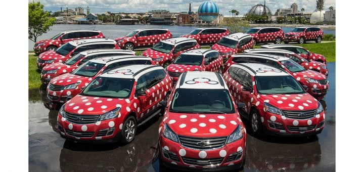 A group of Disney World Minnie Vans, featuring red bodies with white polka dots and Minnie Mouse bows on the roofs, are parked on grass near a waterfront, with buildings and a blue balloon in the background.