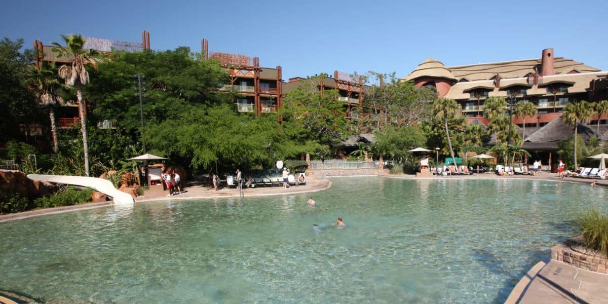 Large outdoor hotel pool at Animal Kingdom Lodge, surrounded by lounge chairs, umbrellas, lush green trees, and a rustic multi-story resort building. A few people swim and relax under clear blue skies.