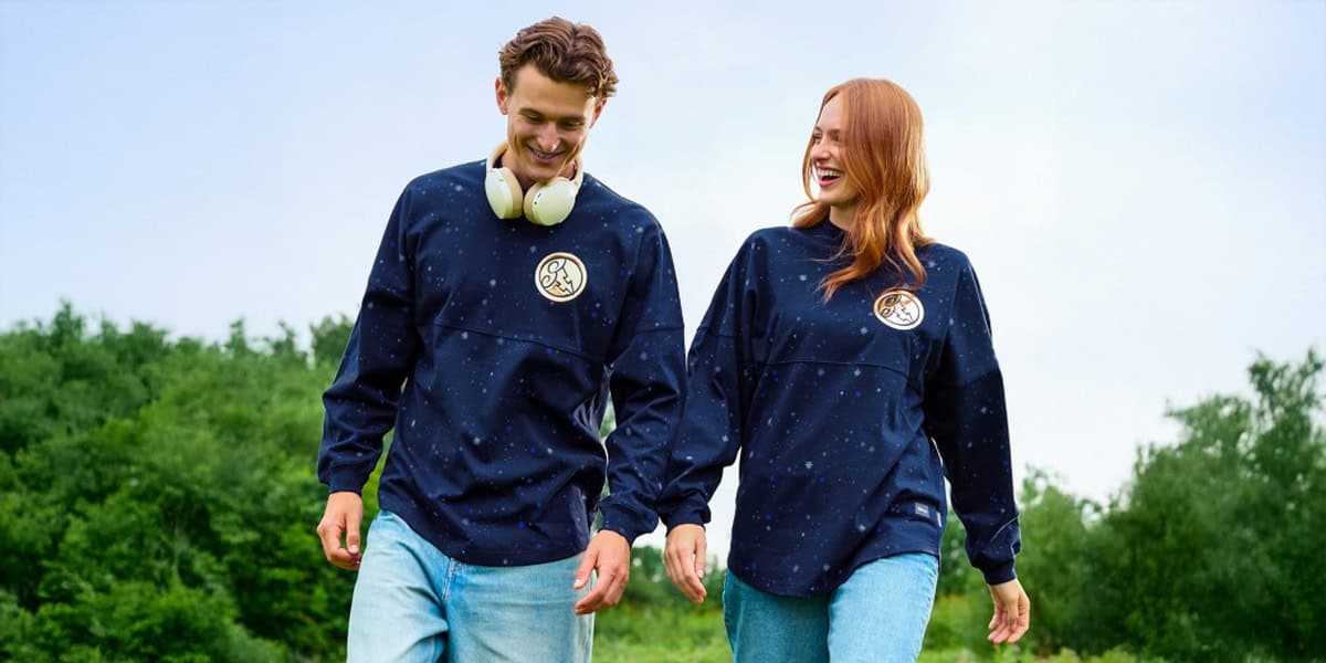 A man and woman stroll outdoors on grass, both smiling and wearing matching navy blue Spirit Jersey sweatshirts with a circular logo—channeling hero vibes. The man has headphones around his neck. Green trees and a blue sky complete the Hercules-worthy scene.