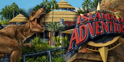 A large T. rex roars near the Jurassic Park area at Universal’s Islands of Adventure, with lush palm trees, round buildings, and the park’s red and yellow sign in the foreground at Islands of Adventure at Universal.