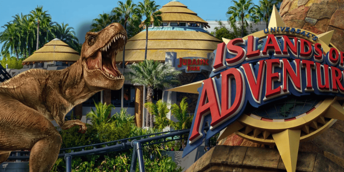 A large T. rex roars near the Jurassic Park area at Universal’s Islands of Adventure, with lush palm trees, round buildings, and the park’s red and yellow sign in the foreground at Islands of Adventure at Universal.