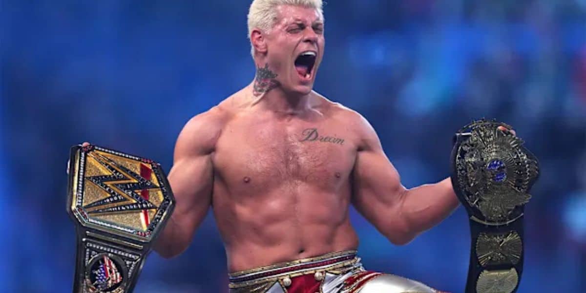 A shirtless, muscular man with blonde hair shouts triumphantly while holding two WWE championship wrestling belts, one in each hand, against a blue-lit arena background.