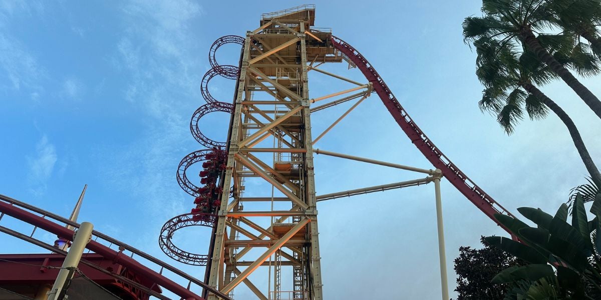 A tall, red and yellow roller coaster features a steep vertical drop and multiple loops, set against a blue sky with a few palm trees and greenery visible on the sides.