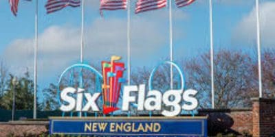 Entrance sign for Six Flags New England with the logo and several American flags waving above it, against a backdrop of trees and blue sky.