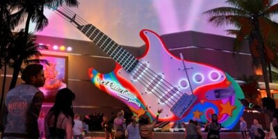 A crowd gathers at night in front of a large, colorful, guitar-shaped entrance to the Rock ‘n’ Roller Coaster attraction, with neon lights, palm trees, and spotlights illuminating the lively scene as The Muppets take over a Disney World ride.