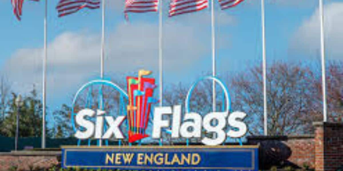 Entrance sign for Six Flags New England with the logo and several American flags waving above it, against a backdrop of trees and blue sky.