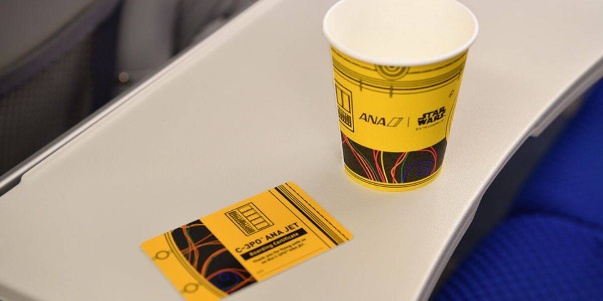 A yellow paper cup and a matching rectangular card with "C-3PO ANA JET" branding sit on a light gray airplane tray table.