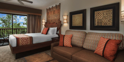 A cozy hotel room at Animal Kingdom Lodge features a large bed, patterned pillows, a brown striped sofa, tribal-inspired wall art, and a balcony overlooking trees and blue sky. Warm lighting creates a welcoming atmosphere.