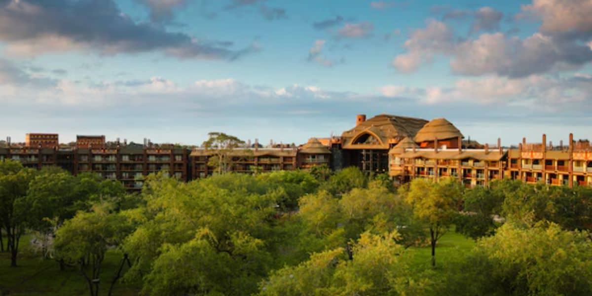A large resort building with an African-inspired design, reminiscent of Animal Kingdom Lodge, stands behind a lush green landscape of trees under a partly cloudy blue sky.