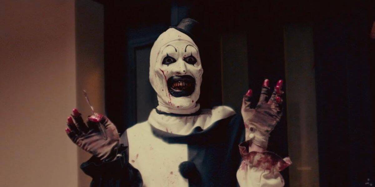 art the clown holds up bloody hands, one holding a small knife while he plasters terrifying grin on his face