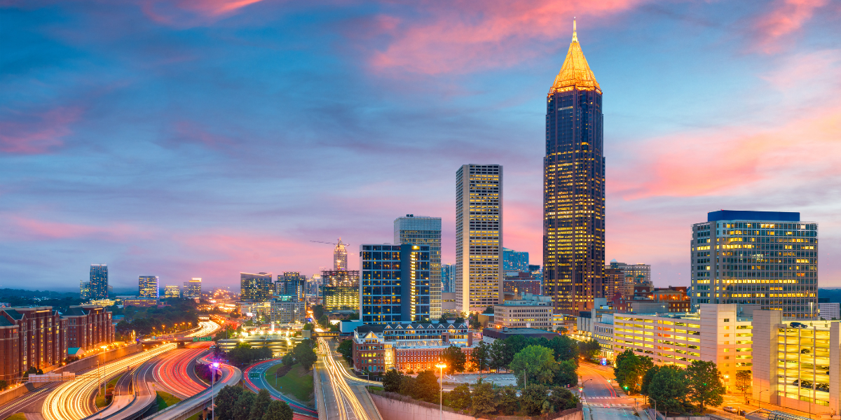 Atlanta, Georgia city skyline at sunset with tall buildings lit up, colorful clouds in the sky, and blurred car lights flowing along highways below.