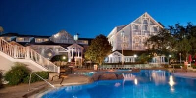 A large, elegant resort hotel with white trim is illuminated at night, reflecting in a spacious, inviting outdoor swimming pool surrounded by trees and lounge areas.