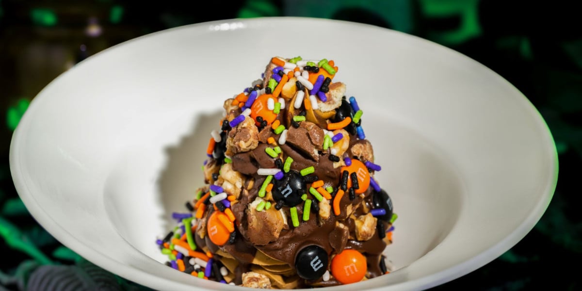 A bowl of chocolate ice cream topped with colorful Halloween-themed sprinkles, popcorn, and orange and black M&M candies, served in a white dish—perfect for adding a touch of Disney magic to your festive treat.