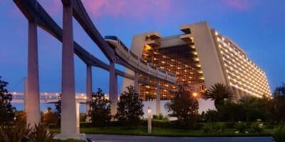 A monorail glides through a modern, A-frame resort hotel at dusk, surrounded by trees and greenery, set against a vibrant purple and blue sky.