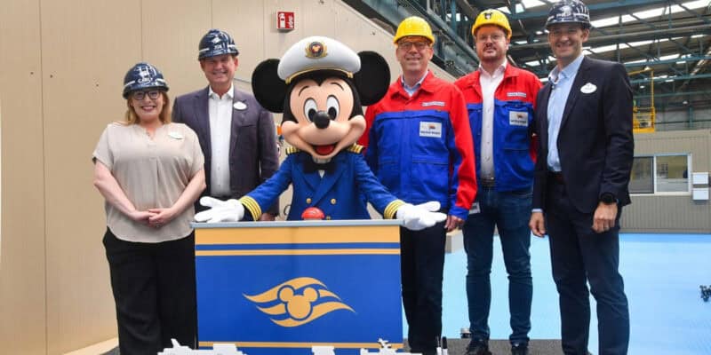 Mickey Mouse in a captain’s uniform stands at a podium with five adults in hard hats and business or work attire inside an industrial building, all smiling during a Disney Cruise Line Steel-Cutting Ceremony.