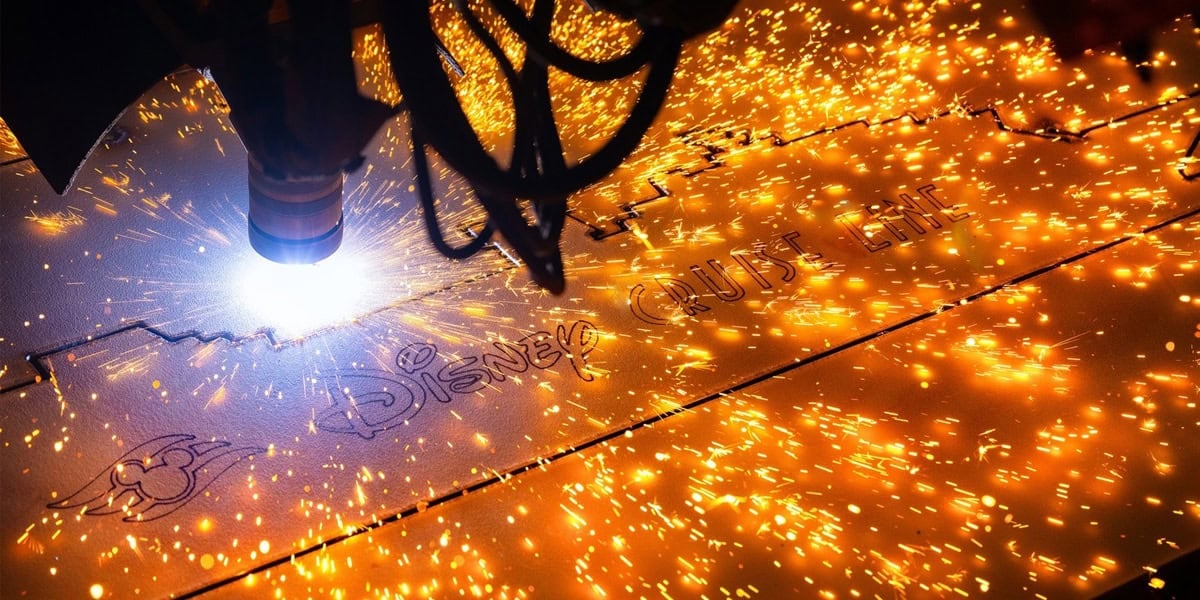 A plasma cutter slices through metal, creating bright orange sparks during a steel-cutting ceremony, as the words "Disney Cruise Line" and a logo are engraved onto the surface.