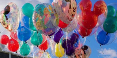 A bunch of colorful balloons decorated with Disney characters, including Mickey Mouse faces and Disney princesses, float against a bright blue sky—capturing the cheerful magic untouched by recent Disney changes for unruly guests.