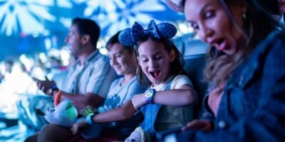 A young girl wearing Mickey Mouse ears excitedly looks at her MagicBand, sitting next to two adults and another child in a lively, colorful indoor Disney World setting.