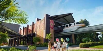 A family walks outside a tropical-themed building with colorful geometric patterns at one of Disney’s Most Expensive Resorts as a monorail passes overhead, surrounded by lush greenery under a blue sky.