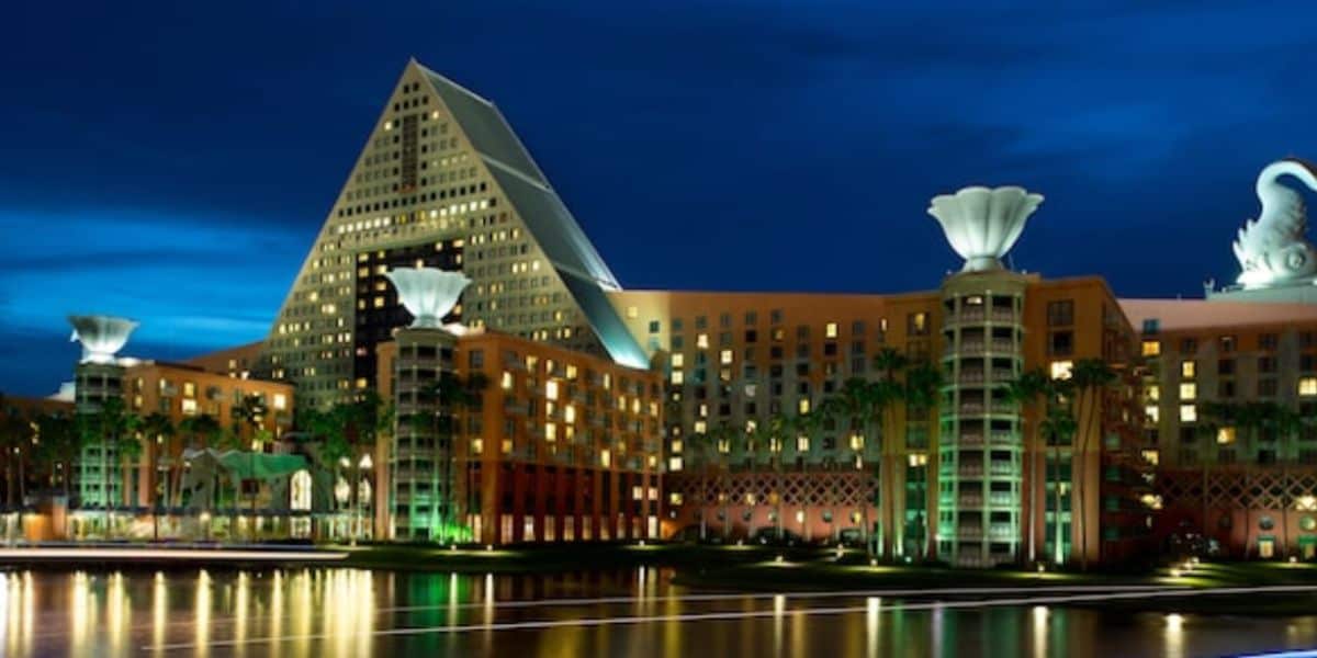 A large, illuminated hotel near Magic Kingdom features a triangular central tower and two tall decorative columns, standing by a calm, reflective body of water at night under a deep blue sky.