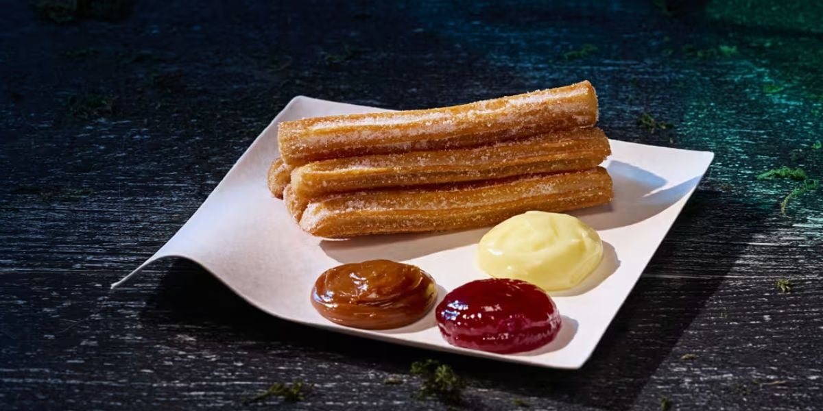 A white plate holds four sugar-coated churros and three dipping sauces—caramel, strawberry, and vanilla—arranged neatly on a dark wooden surface, making the perfect treat to enjoy after thrilling horror nights.