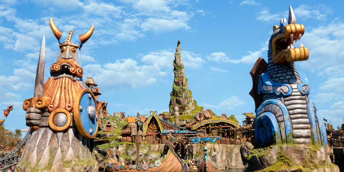 Colorful amusement park scene with a large Viking warrior statue holding an axe and shield on the left, a blue dragon-like statue on the right, themed buildings, a rocky mountain, and a playful Donkey Kong figure in the background at Epic Universe.