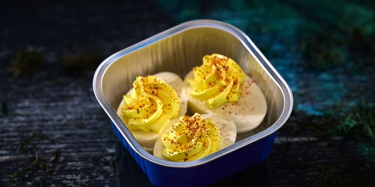 Three deviled eggs topped with yellow yolk filling and sprinkled with paprika are arranged in a small, rectangular metal container on a dark surface—perfect for snacking during horror nights.