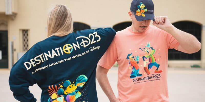 Two people sport exclusive finds from Disney’s “Destination D23 2025.” One shows off a navy sweatshirt with Disney characters, while the other wears a peach t-shirt and navy cap, both featuring stylish Mickey Mouse designs.