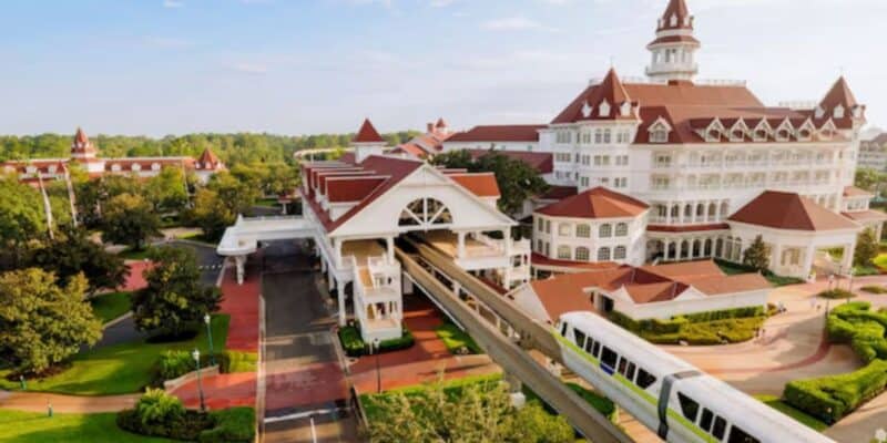 A white monorail glides past a large, elegant resort hotel with red roofs and white walls, surrounded by greenery and pathways under a sunny sky.