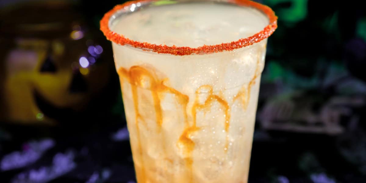A clear plastic cup filled with a light, bubbly drink, drizzled with caramel and topped with a red sugar rim, sits in front of a blurred dark background featuring a faint Disney Halloween pumpkin decoration.