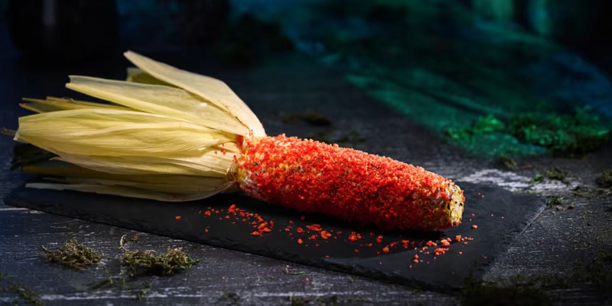 A corn on the cob with husks partially peeled back, coated in a bright red seasoning, sits on a dark slate surface with a moody background and green moss-like decorations—perfect for horror nights-inspired gatherings.