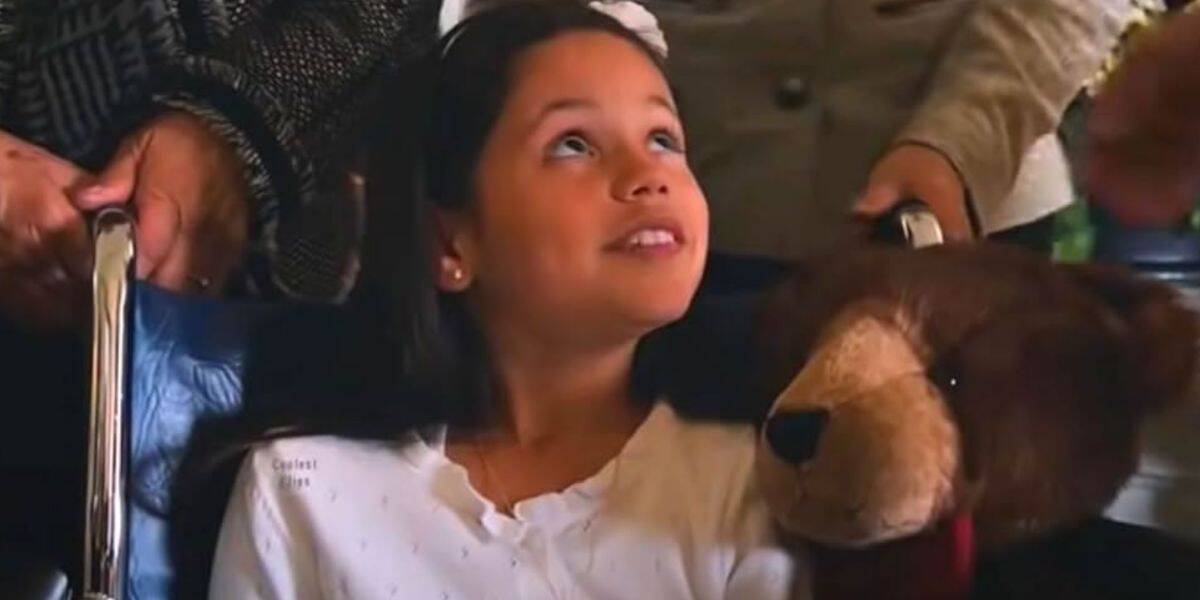 A young girl with long dark hair, reminiscent of Jenna Ortega, sits in a wheelchair with a white bow, smiling up as she holds a plush brown bear. Adults gently rest their hands on the wheelchair around her.