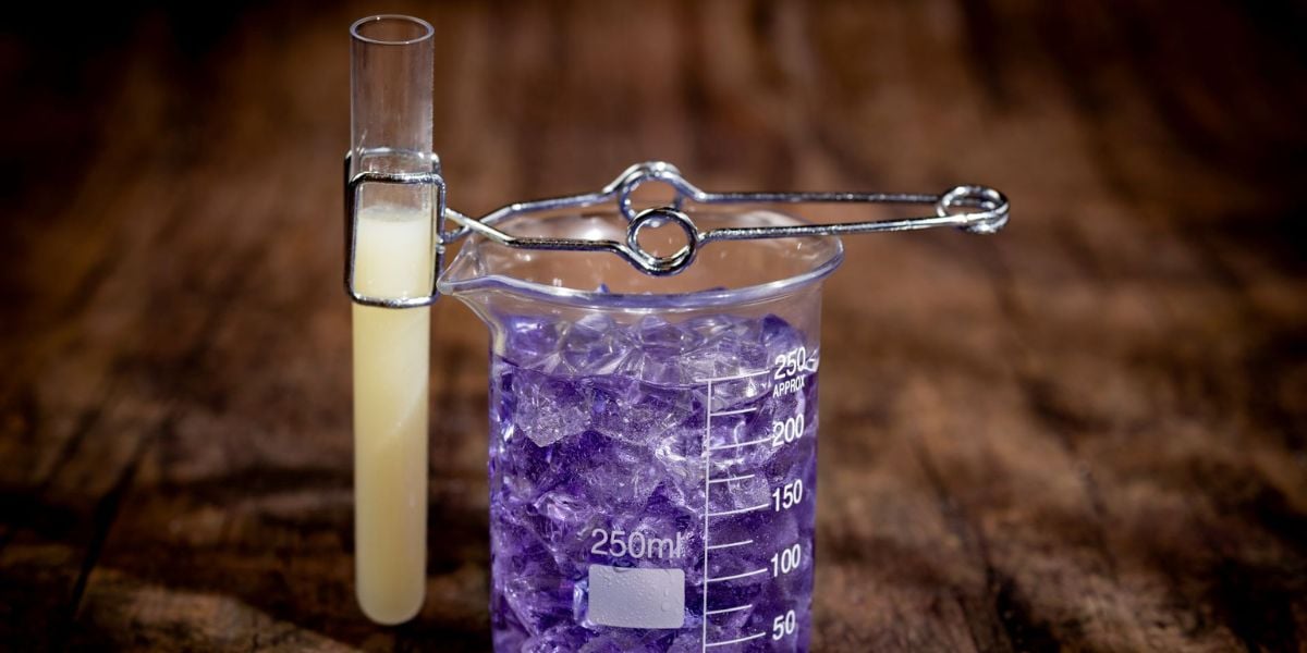 A test tube with a cloudy yellow liquid is held by tongs above a 250 ml beaker filled with purple liquid and ice cubes, both set on a wooden surface—like planning Disney World Holiday Pricing, every detail matters.