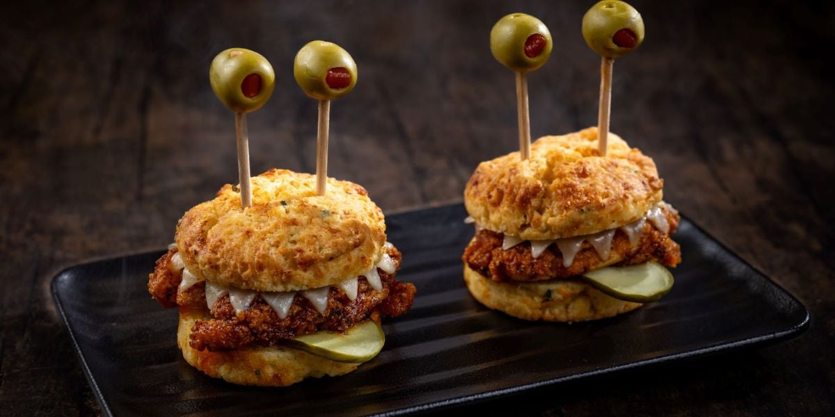 Two chicken biscuit sandwiches topped with pickles and cheese, each with two toothpicks holding green olives as "eyes," are served on a black rectangular plate—perfect for a festive Disney World Holiday Pricing brunch.
