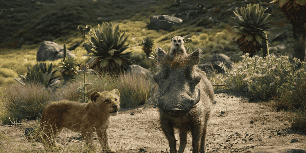 A young lion, a warthog, and a meerkat stand together in a sunlit, grassy landscape with rocks and lush plants, ready for new adventures—like those seen in animated sequels. The meerkat is perched on the warthog’s head, all facing forward.