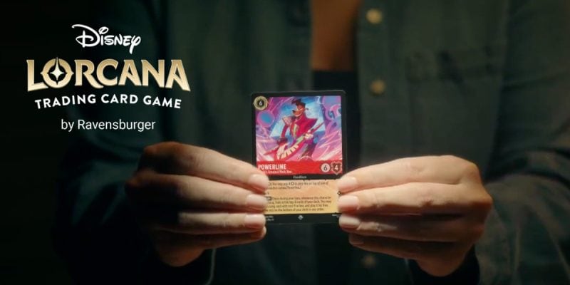 A person holds a Disney Lorcana trading card featuring Powerline, celebrating the second anniversary of Lorcana. The Disney Lorcana logo and "Trading Card Game by Ravensburger" text appear on the left side of the image.
