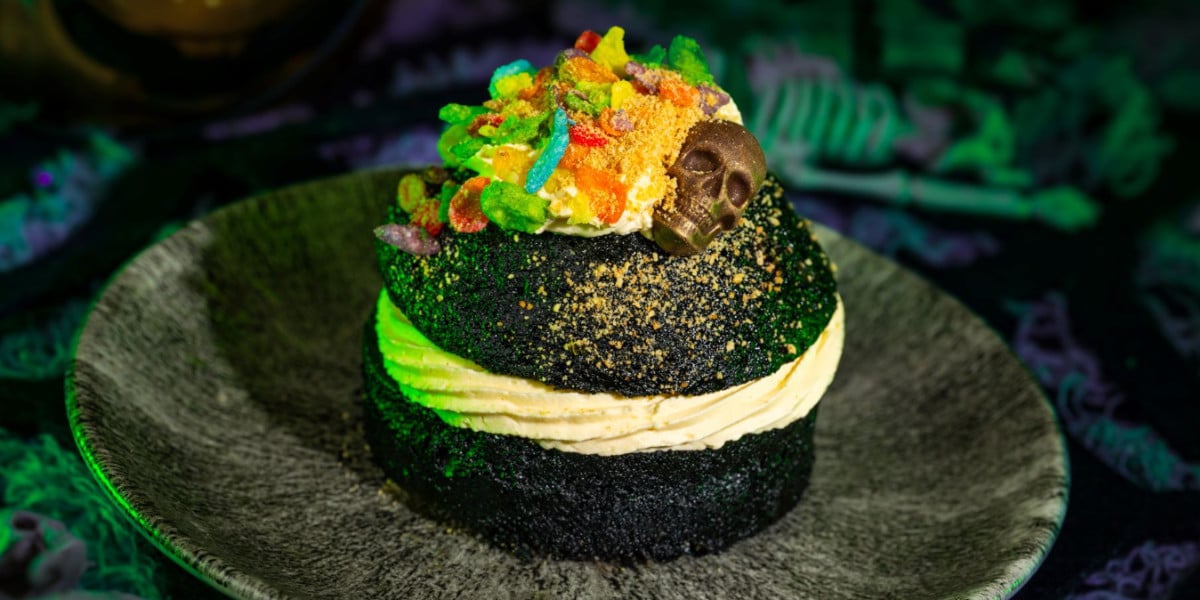A dark chocolate cake with cream filling, topped with colorful cereal bits, a swirl of frosting, and a small chocolate skull, served on a dark plate with a greenish glow—perfect for fans of Disney’s delightfully spooky treats.