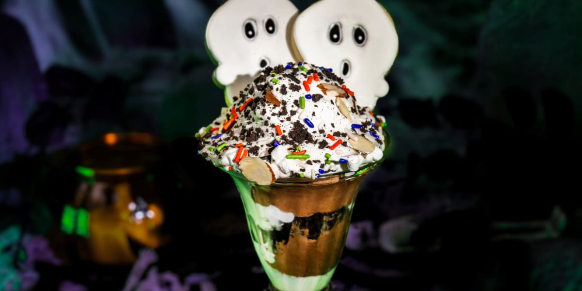 A Disney-inspired Halloween ice cream sundae in a glass, topped with whipped cream, chocolate, colorful sprinkles, crushed cookies, and two white ghost-shaped cookies with black eyes.