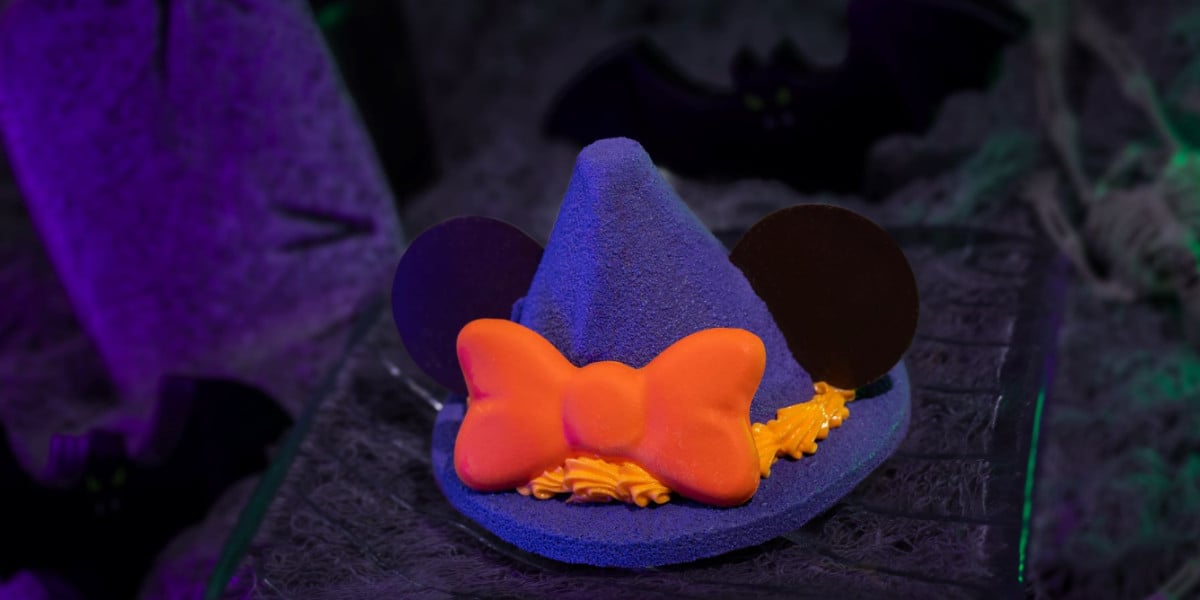 A Disney-inspired dessert shaped like a purple witch hat with chocolate mouse ears and an orange bow, set on a dark, Halloween-themed background with bats and dim lighting.