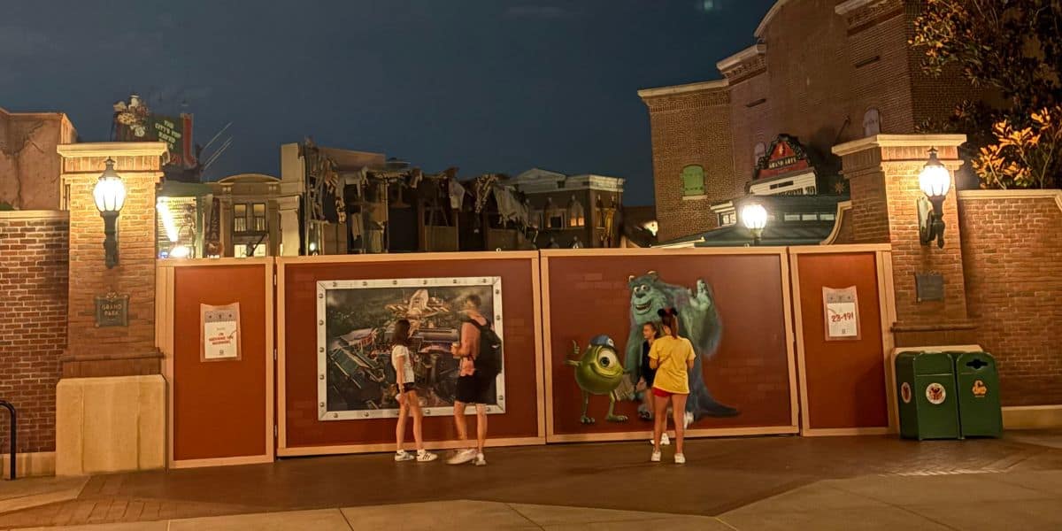 Two people stand in front of construction walls at night featuring Pixar’s Monsters, Inc. art. Another person in yellow faces the wall; buildings and street lamps glow behind them, hinting at changes like when Disney gets rid of Muppets.