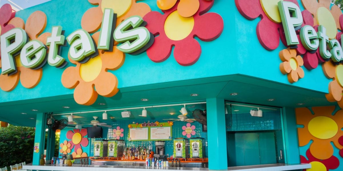A brightly colored bar called Petals, inspired by pop century style, features large, vibrant flower shapes on a turquoise wall, with drinks and menus visible at the open counter.