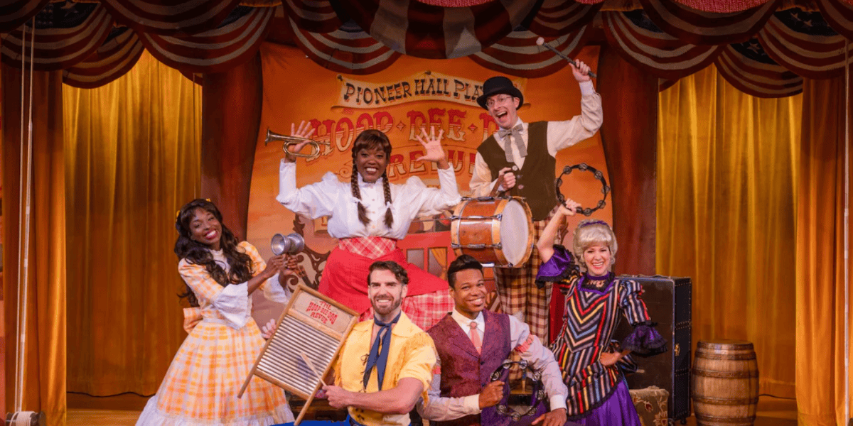 Six performers in colorful, old-fashioned costumes pose on stage with musical instruments in front of a “Pioneer Hall” banner at Fort Wilderness. The backdrop and decor evoke a lively, Western-themed show.