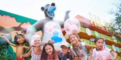 Five smiling children wearing Disney-themed hats and mouse ears pose in front of large Baloo and Mowgli statues at Pop Century, a colorful theme park with balloons and cheerful decorations visible.