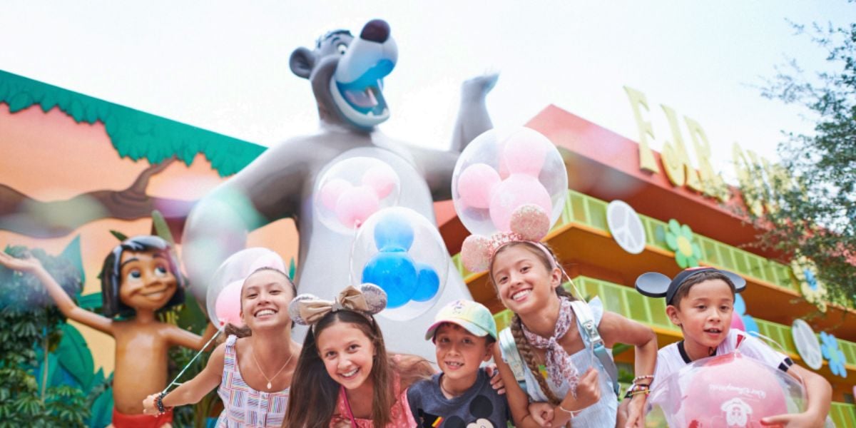 Five smiling children wearing Disney-themed hats and mouse ears pose in front of large Baloo and Mowgli statues at Pop Century, a colorful theme park with balloons and cheerful decorations visible.