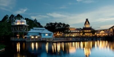 A waterfront resort view at dusk shows buildings with warm lights reflecting on the calm water, surrounded by trees. A water tower and a dock are visible on the left, with more illuminated buildings in the background at this Disney World hotel.