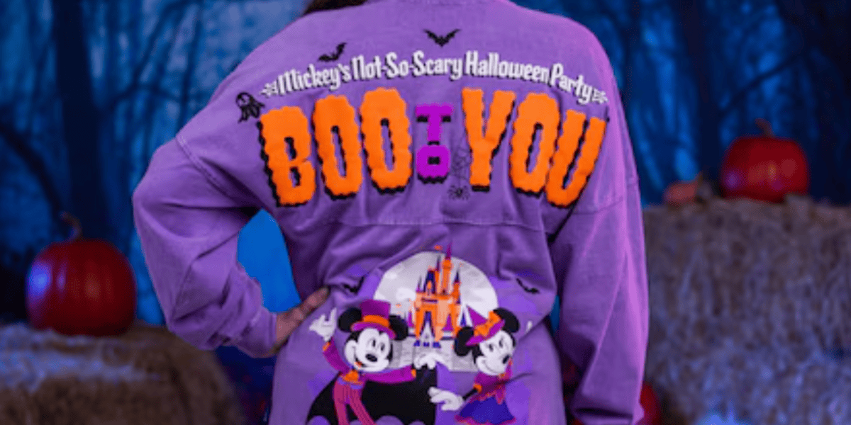 A person wearing a purple Halloween-themed jacket with "Mickey's Not-So-Scary Halloween Party BOO TO YOU" and images of Mickey and Minnie Mouse in costumes, standing in front of hay bales and pumpkins at a festive halloween party.