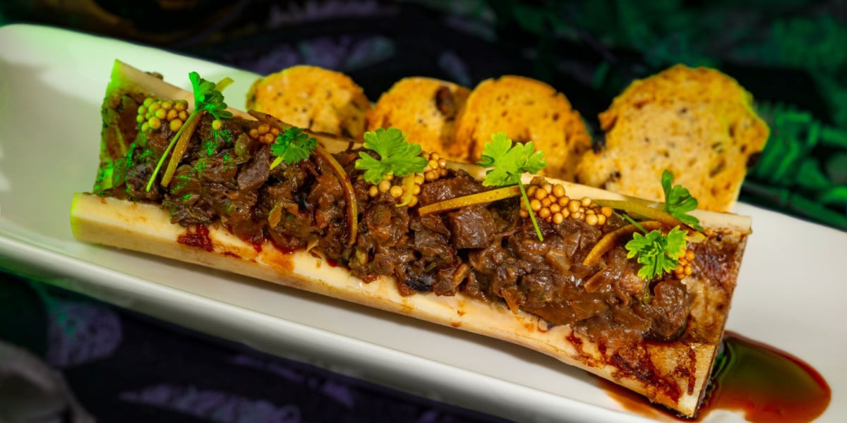 A roasted bone marrow half filled with a savory meat mixture, garnished with herbs and pickled mustard seeds, sits on a white plate—like a dish from a Disney feast—with three slices of toasted bread in the background.