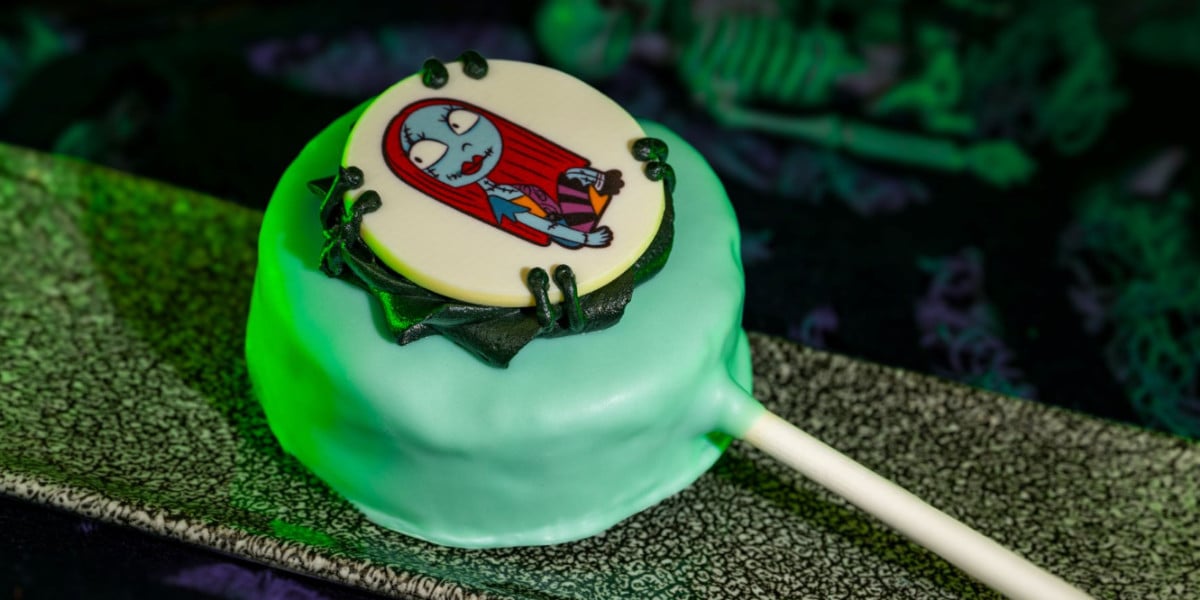 A Disney cake pop coated in light blue icing features an edible image of Sally from "The Nightmare Before Christmas." The treat sits on a dark rectangular plate with a lollipop stick, surrounded by Halloween-themed decorations.