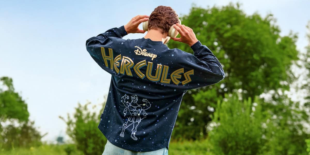 A person standing outdoors, seen from behind, wears a navy blue Disney Hercules Spirit Jersey featuring gold lettering and an illustration of the hero himself, with trees and greenery in the background.