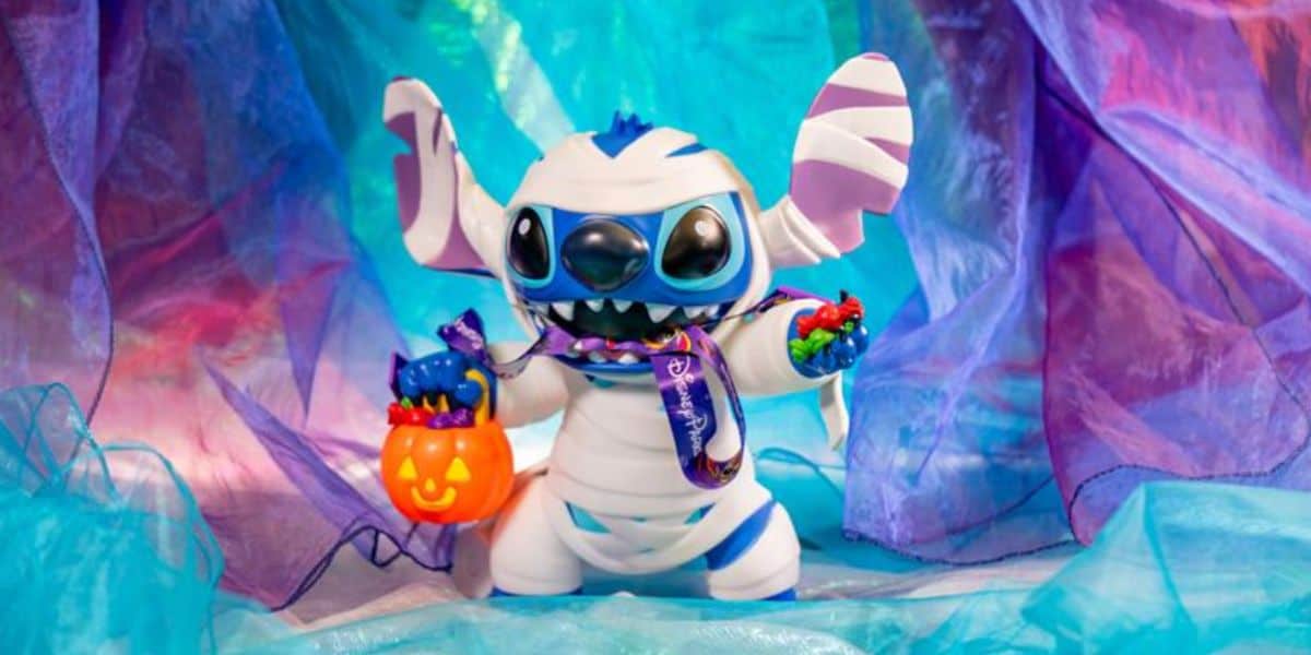 A Stitch figurine dressed as a mummy holds a Halloween pumpkin bucket filled with colorful candies, surrounded by vibrant blue and purple sheer fabric—perfect for any Stitch fan’s spooky season display.